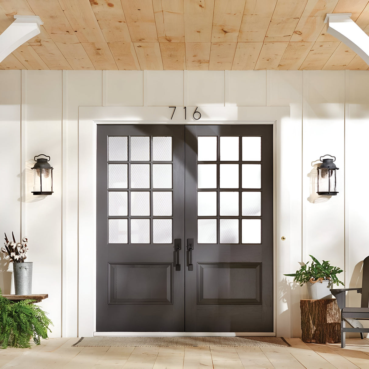 A set of black double doors with square glass panes is centered on a white-paneled exterior wall, with the number 716 displayed above. Flanking the doors are two Ashland Bay Outdoor 1 Light Large Wall Lanterns in Weathered Zinc, fitted with LED Edison bulbs, while a small arrangement of plants sits beside one lantern.