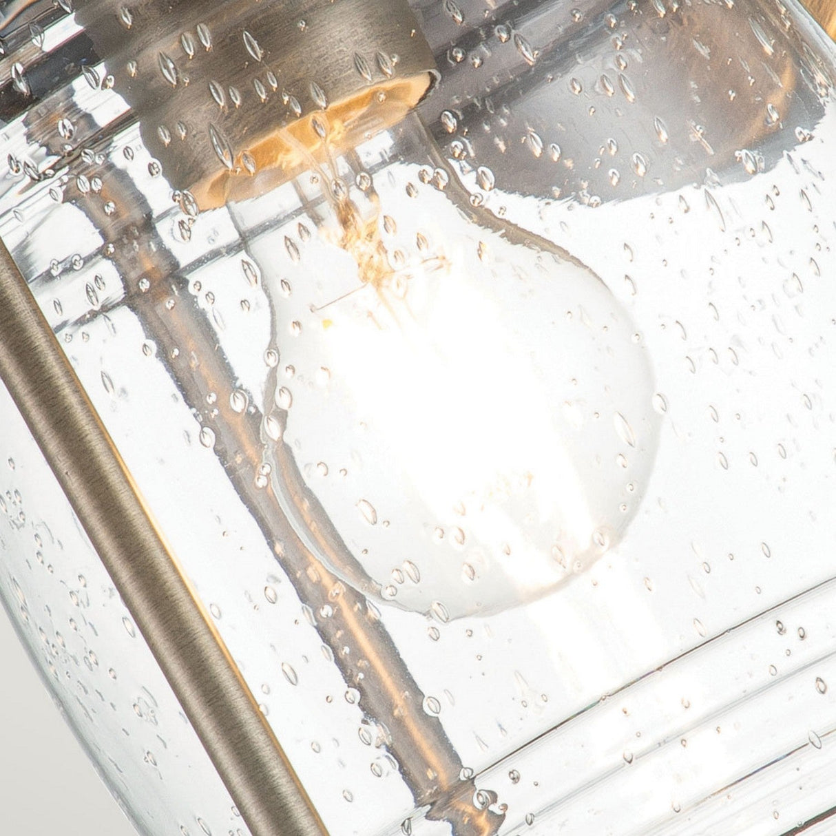 A close-up of the Ashland Bay Outdoor 1 Light Medium Wall Lantern, featuring a burnished bronze frame, displays a lit bulb within clear seeded glass. The illuminated bulb enhances the speckled design and textured surface against the metal framework.