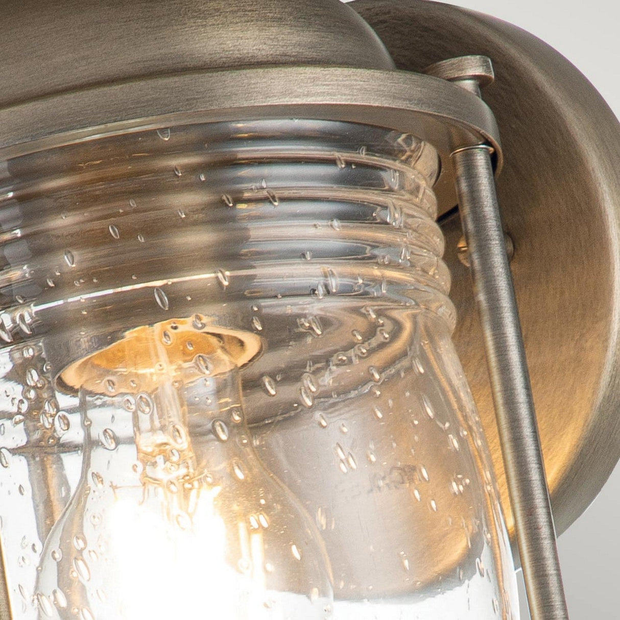 A detailed shot of the Ashland Bay Outdoor 1 Light Small Wall Lantern features a vintage design with a clear seeded glass shade and a warm LED Edison bulb. This fixture, with its burnished bronze finish, is mounted on a curved arm to create an inviting glow.