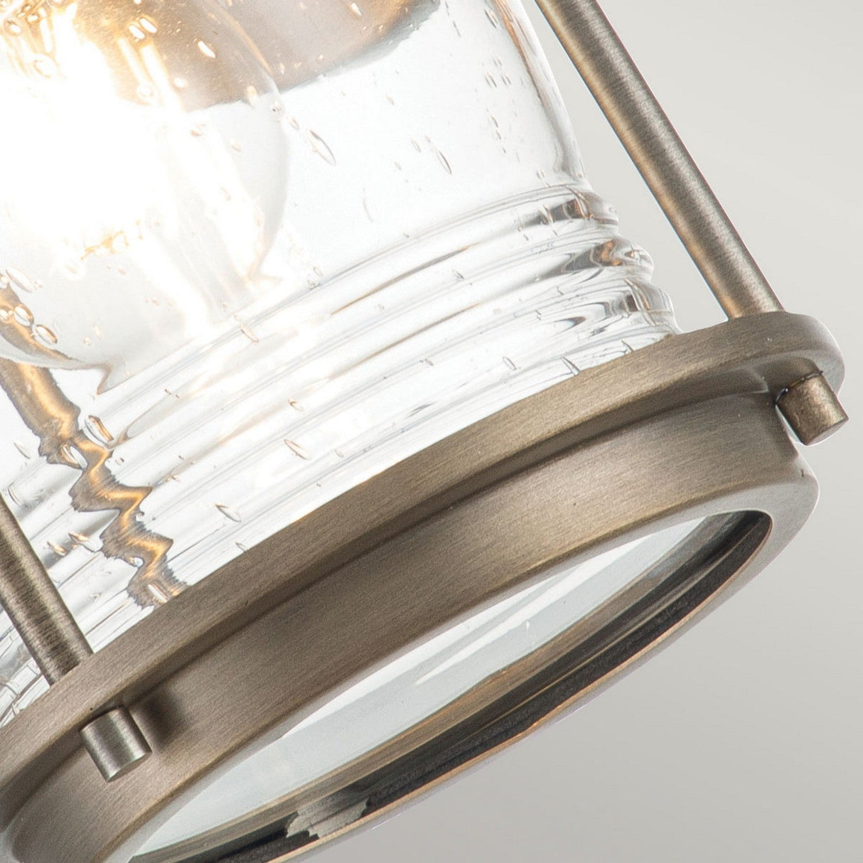 Close-up of the Ashland Bay Outdoor 1 Light Small Wall Lantern in burnished bronze, featuring a clear, textured glass cover. Fitted with LED Edison bulbs, this vintage-style fixture emits a warm glow that accentuates its design details against a neutral background.