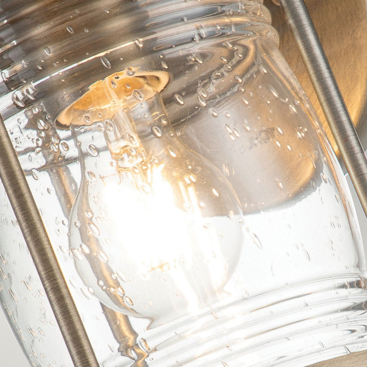 A close-up of the Ashland Bay Outdoor 1 Light Small Wall Lantern in burnished bronze reveals a glowing LED Edison bulb inside a textured clear glass fixture adorned with small water droplet designs. The warm light accentuates the contrast between the glass and metallic elements, showcasing its timeless charm.