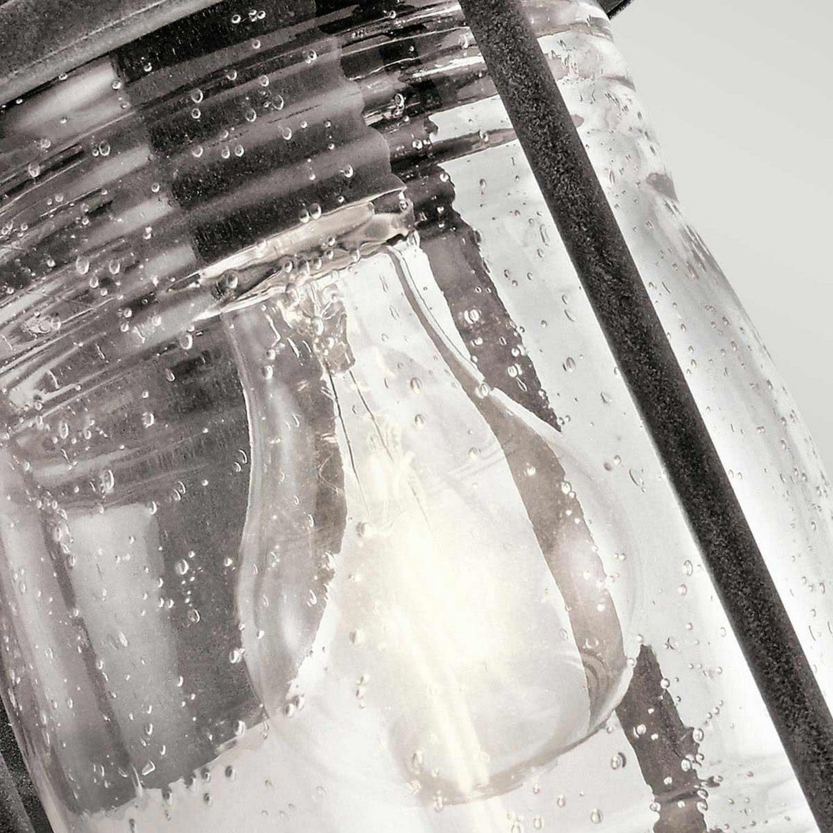 A close-up of the Ashland Bay Outdoor 1 Light Small Wall Lantern reveals a light bulb enclosed within a weathered zinc frame. The glass, speckled with moisture droplets, adds a textured effect that evokes a timeless colonial design. Its rustic charm is enhanced by the soft, out-of-focus gray background.