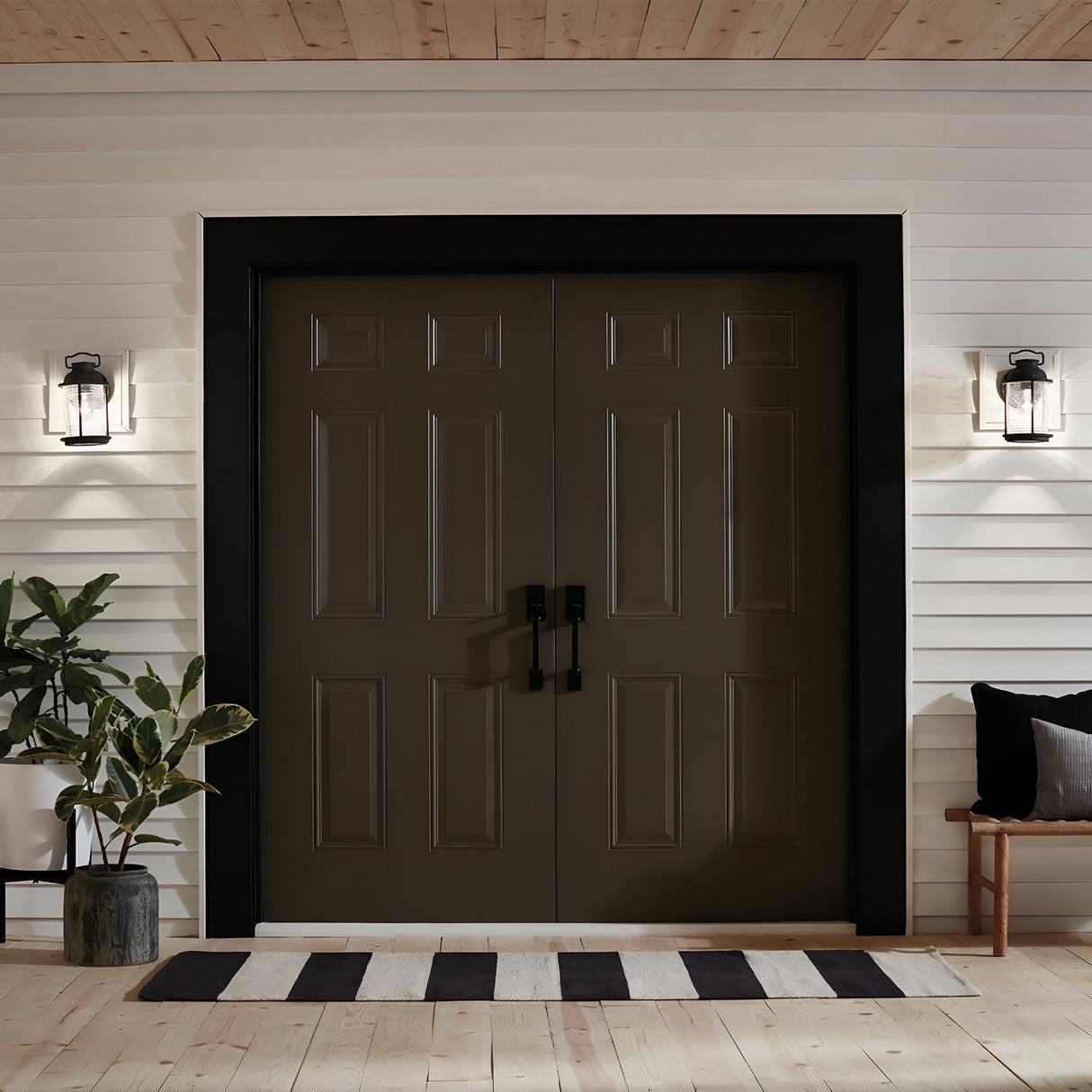 A modern entryway features double dark brown doors with black trim, complemented by Ashland Bay Outdoor 1 Light Small Wall Lanterns in a sleek Colonial Design. On one side rests a small plant, while a wooden bench with cushions occupies the other. A striped rug in front adds warmth to the space.