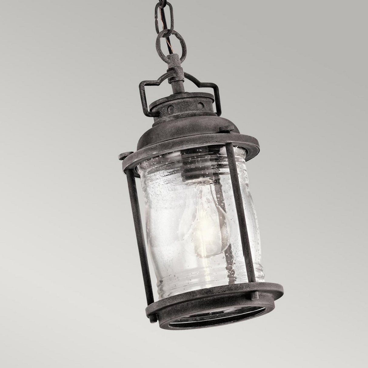 Introducing the Ashland Bay Outdoor 1 Light Small Chain Lantern in Weathered Zinc, a vintage-inspired piece with rustic metal and glass elements. This cylindrical lantern features clear glass panels and an Edison-style LED bulb inside, all suspended by a chain against a plain, light gray backdrop.