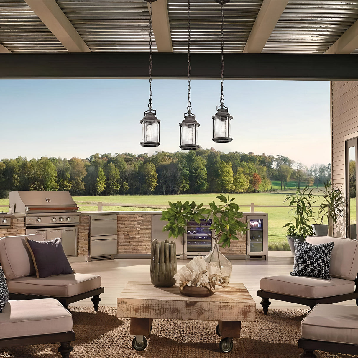 A spacious outdoor kitchen and lounge area is equipped with a grill, mini-fridges, and a weathered zinc wooden table adorned with plants. Overhead, an Ashland Bay Outdoor Chain Lantern in Weathered Zinc casts light onto the scene. The area offers comfortable seating around the table, all set against a backdrop of open fields and trees.