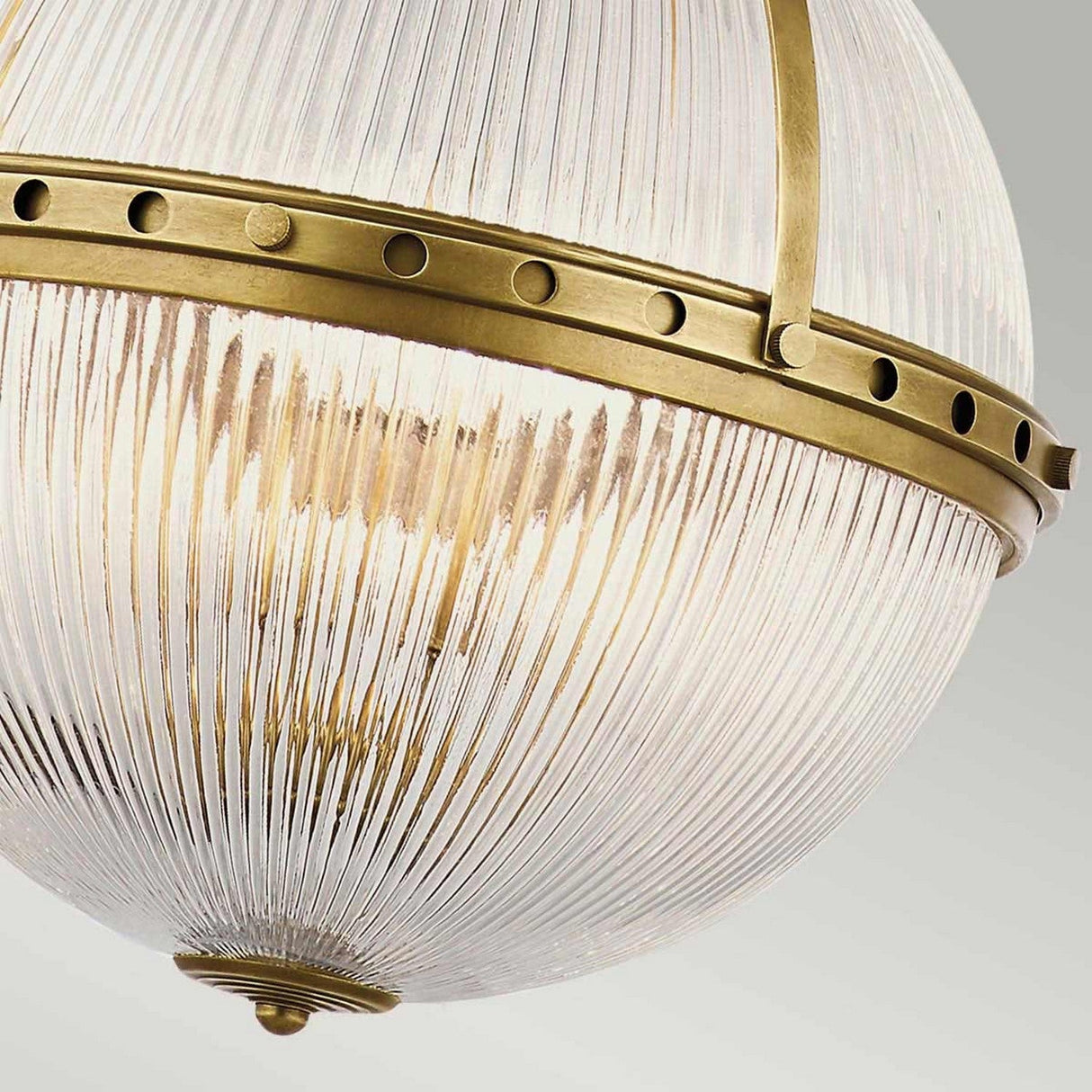 A close-up of the Aster 3 Light Pendant - Brass showcases vintage-style ribbed glass domes, accented by a brass band with rivet details encircling the middle. The image captures the warm glow of the light within, highlighting the intricate glass design.