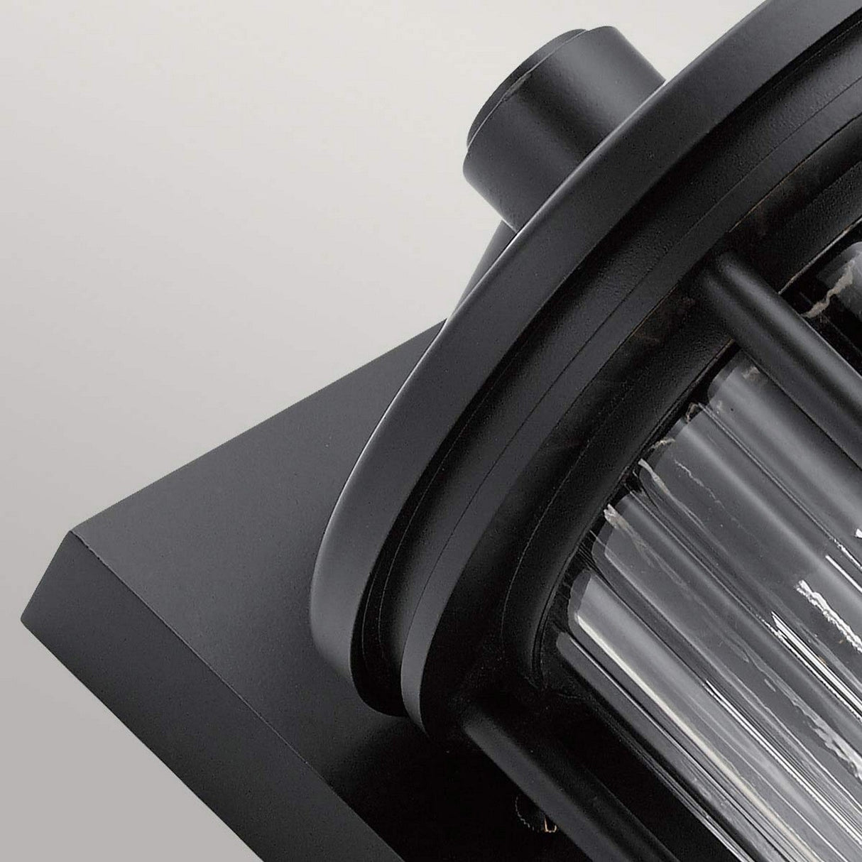 Close-up of the Barras Outdoor 1 Light Medium Wall Lantern in black, showcasing its modern cylindrical design with ribbed glass rods that diffuse light. Mounted on a dark square base against a light gray backdrop, this fixture adds an elegant touch to any outdoor lighting arrangement.