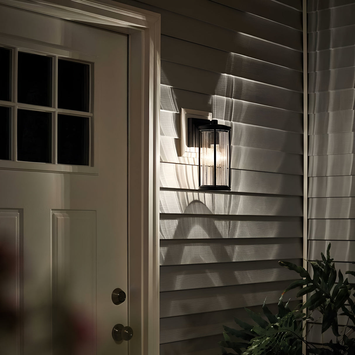 The Barras Outdoor 1 Light Small Wall Lantern in black adds a touch of vintage charm to the siding of a house next to a door with glass panes. Its black finish casts intricate shadow patterns on the wall, while a plant is partially visible in the foreground, completing this serene nighttime scene.