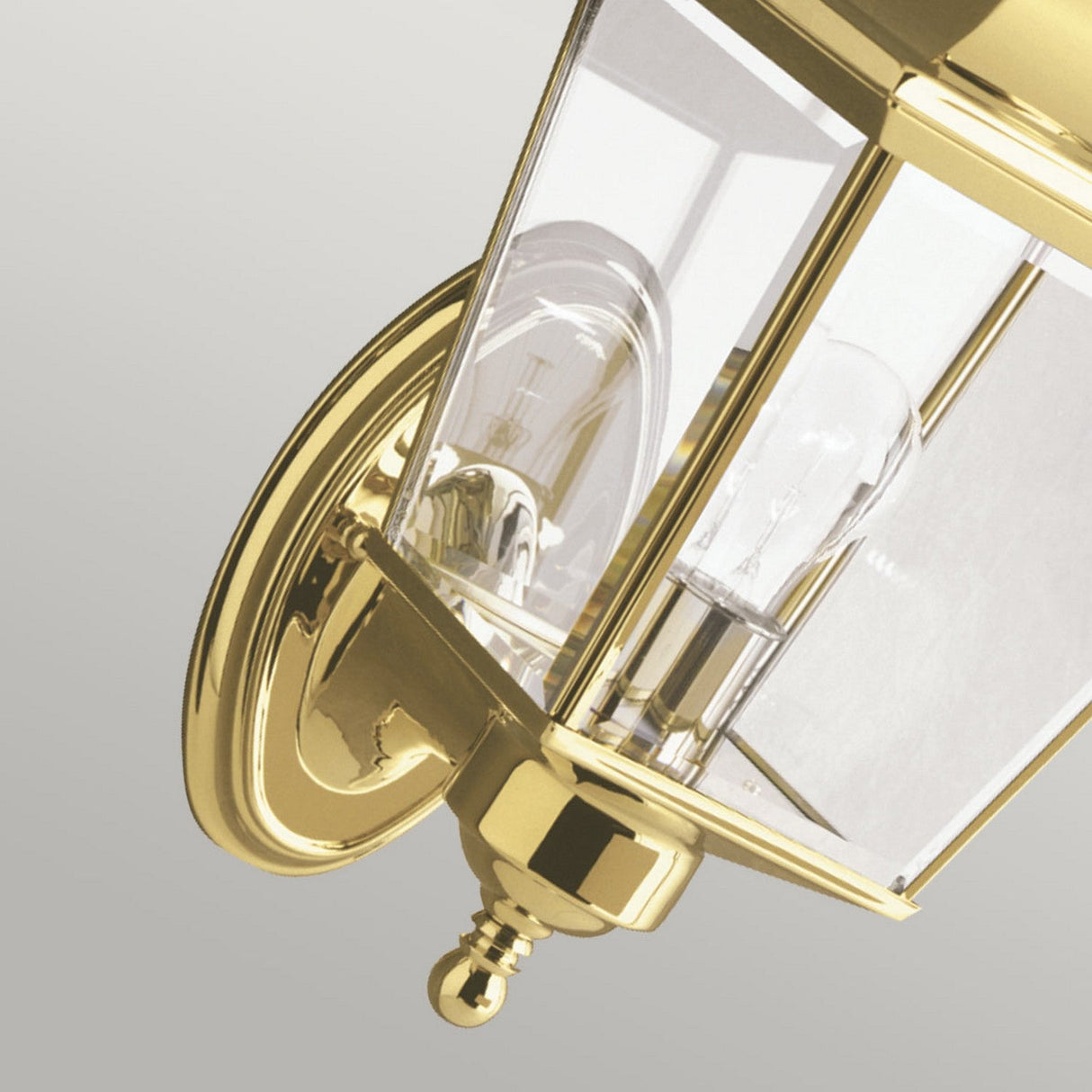 A close-up of the Bay Shore Outdoor 1 Light Medium Wall Light in polished brass showcases its traditional design. The fixture features clear beveled glass panels and an exposed light bulb, all mounted on a circular brass backing. Its elegance is emphasized against a simple gray background.