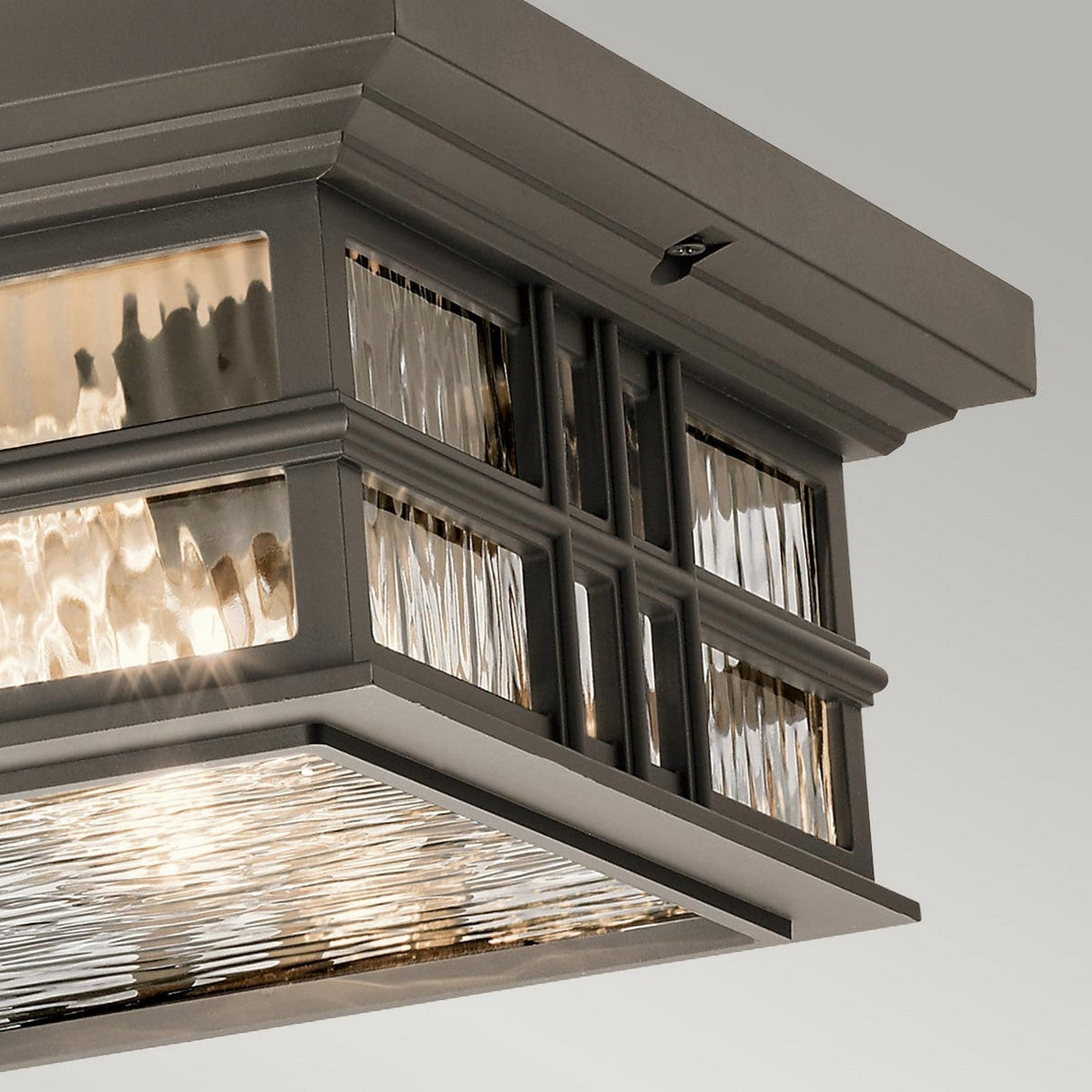 Close-up of the Beacon Outdoor 2 Light Flush - Bronze, featuring a modern rectangular ceiling light fixture in an Olde Bronze finish with textured glass panels and a metallic frame. Its warm glow enhances the reflective surfaces and intricate design, capturing elements of craftsmen architecture reminiscent of Beacon Square outdoor lighting.