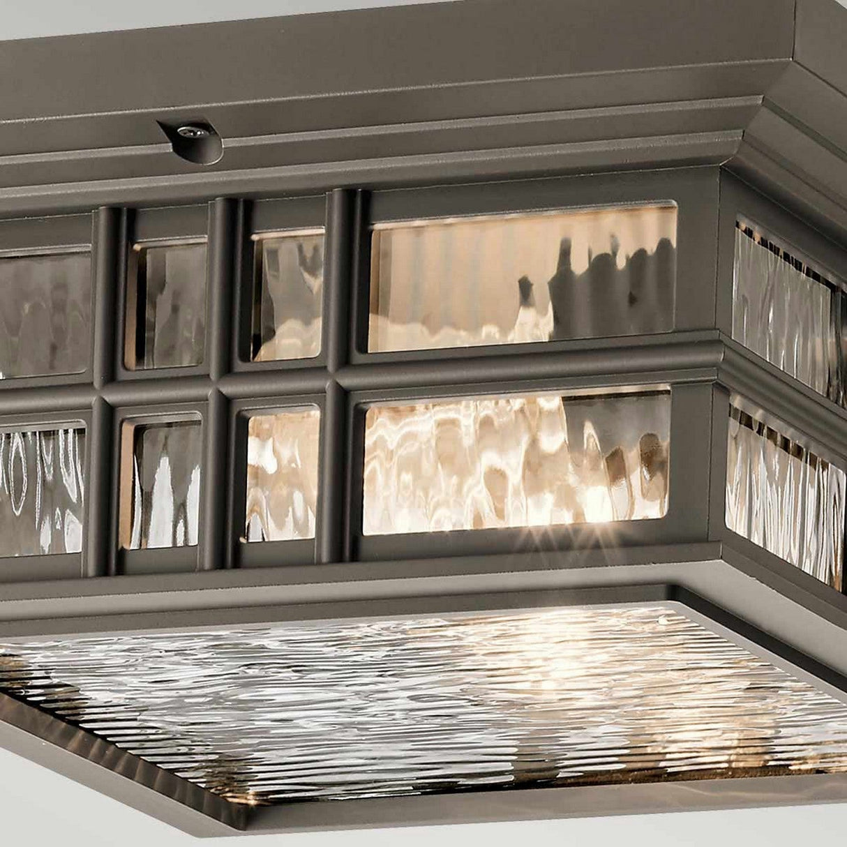 A detailed view of the Beacon Outdoor 2 Light Flush - Bronze showcases its Olde Bronze finish with a brushed metal frame, complemented by textured glass panels that form a wavy pattern. This fixture casts a warm glow through the glass, evoking the essence of craftsman architecture and elegantly highlighting its intricate design.