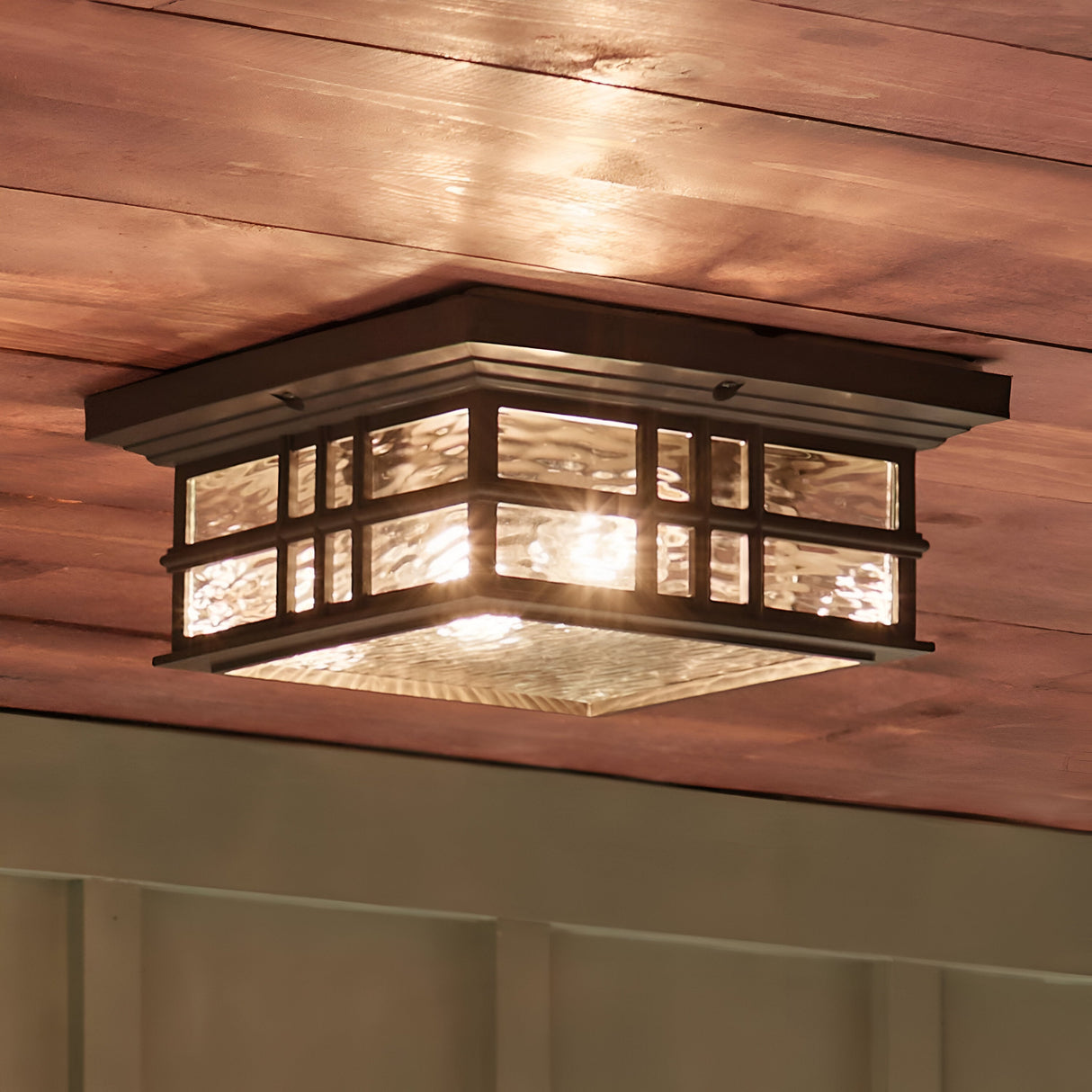 The Beacon Outdoor 2 Light Flush - Bronze from the Beacon Square outdoor collection has a stunning Olde Bronze finish and clear hammered glass panels, perfect for mounting on a wooden ceiling to cast a warm, inviting glow that beautifully enhances any space.