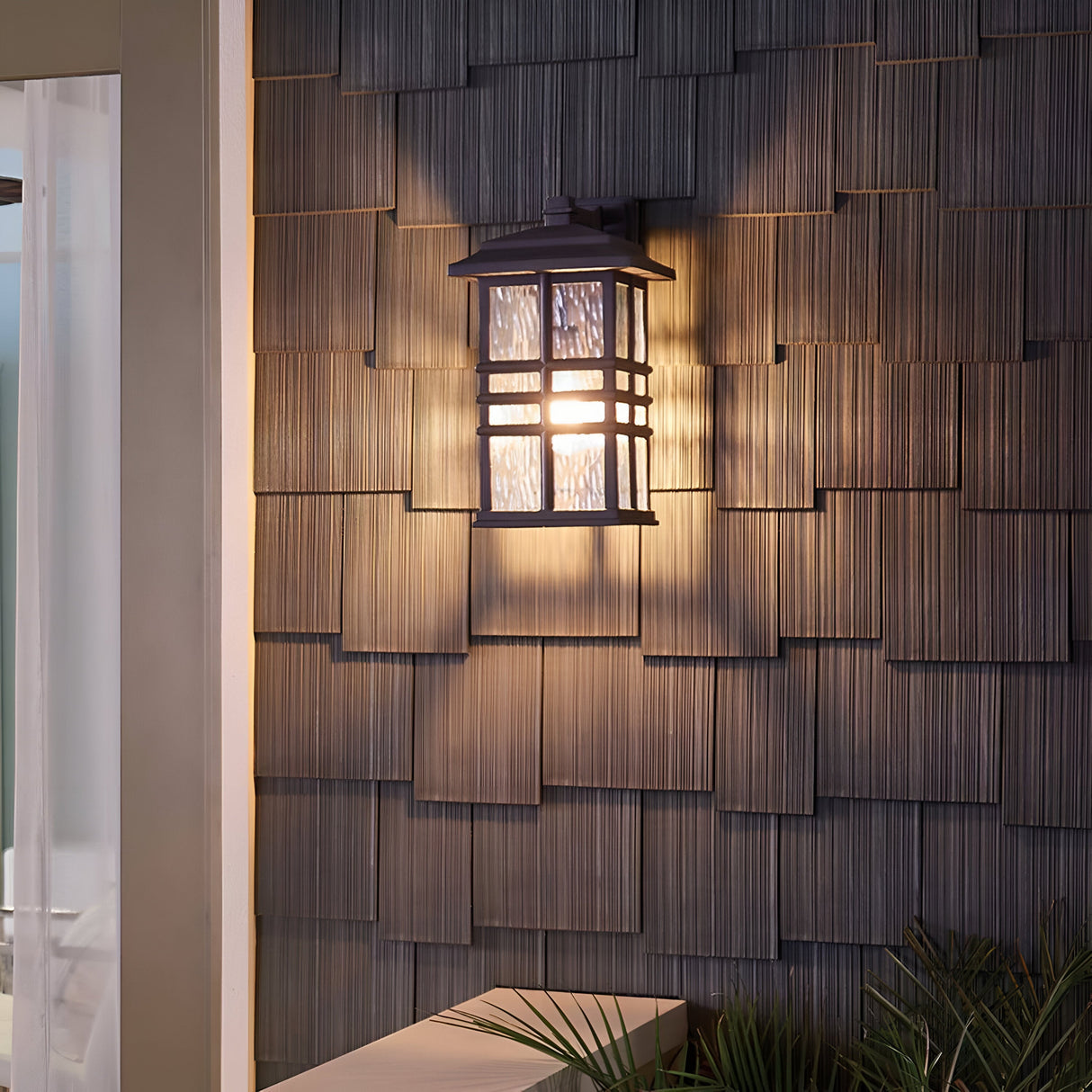 The Beacon Square Outdoor 1 Light Medium Wall Lantern in bronze is mounted on a textured gray wall, casting warm light that creates a cozy ambiance. A small plant is visible at the bottom of the image, while part of a white doorway appears on the left, ensuring it's IP44 rated for durability.