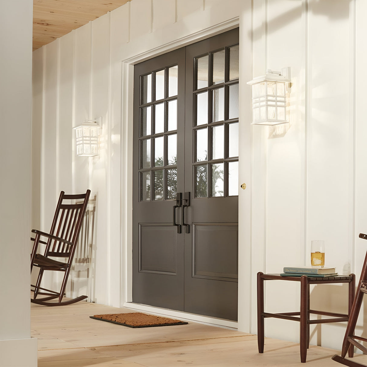 A cozy porch boasts two dark brown wooden rocking chairs and a small table with a glass of drink next to double black doors with windowpanes. The area, designed in the Craftsman style, is illuminated by two Beacon Square Outdoor 1 Light Medium Wall Lanterns in white, while a welcoming doormat lies in front of the doors.