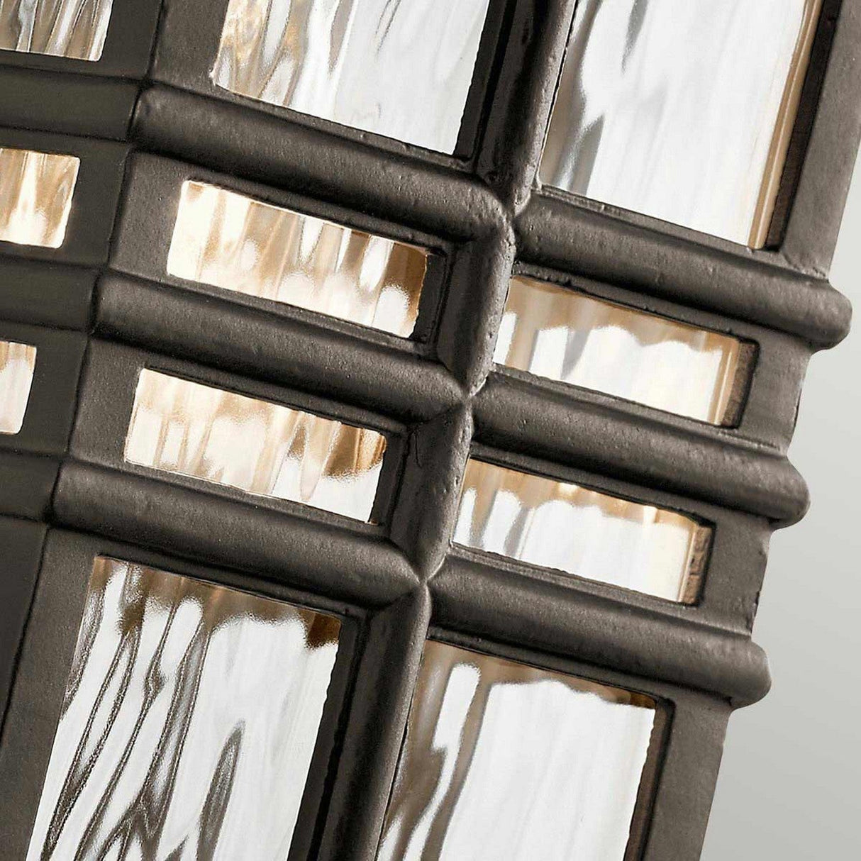Close-up of the Beacon Square Outdoor 1 Light Small Wall Lantern in Bronze, showcasing its decorative glass and metal design with intersecting black metal lines forming a grid pattern. The textured glass panels reflect light, creating a dynamic play of light and shadow reminiscent of a modern, geometric style outdoor lighting fixture.