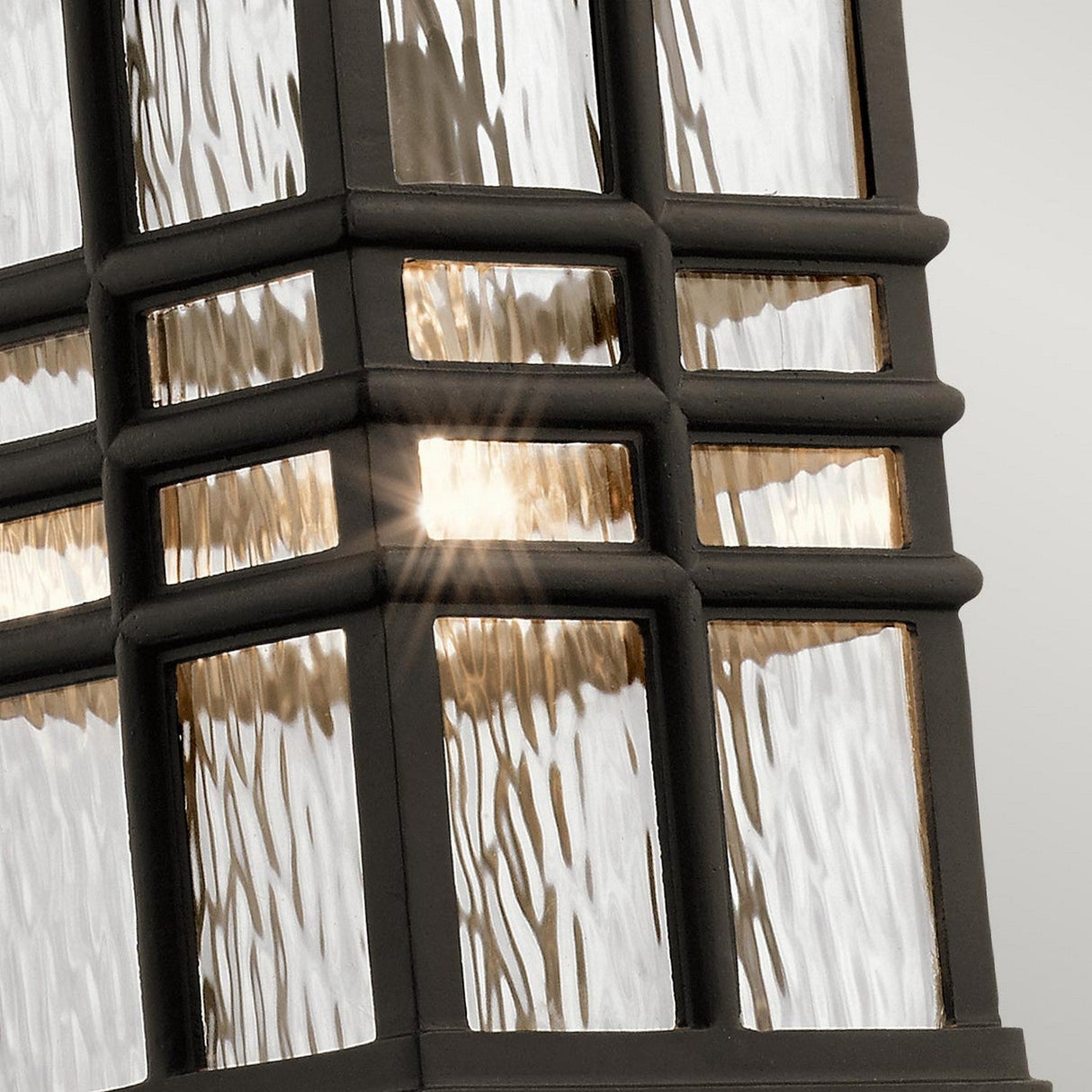 Close-up of the Beacon Square Outdoor 1 Light Chain Lantern in bronze, showcasing its textured glass panes set within a black metal frame. As light shines through, it creates a glowing effect. The lantern's design boasts a geometric pattern with intersecting lines that embodies transitional style lighting.