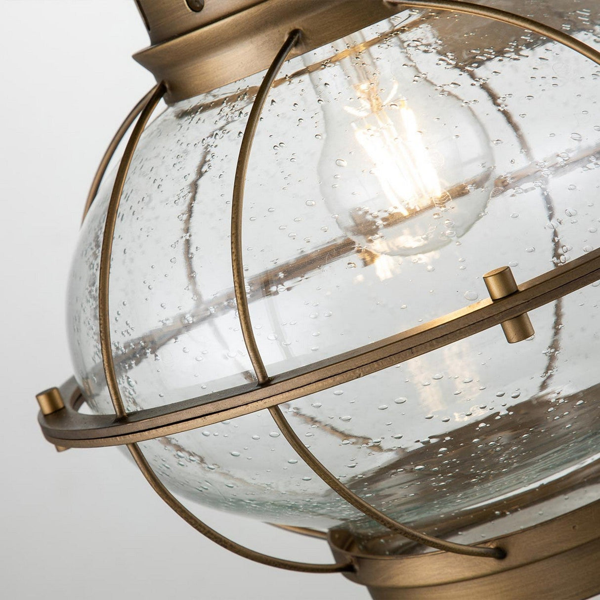 A close-up of the Bridgepoint Outdoor 1 Light Large Wall Lantern in natural brass, featuring a clear, bubbled glass globe with a visible light bulb. The fixture's design includes a metal cage giving it an industrial touch inspired by nautical lighting.