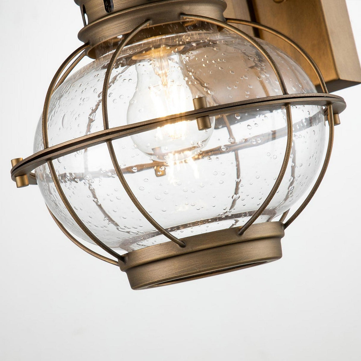 The Bridgepoint Outdoor 1 Light Medium Wall Lantern - Natural Brass showcases a nautical-style design, featuring a natural brass finish and a clear, bubbled glass globe encased in a metal cage. The central light bulb is visible, emitting a warm glow, as the fixture is elegantly mounted on a rectangular backplate.