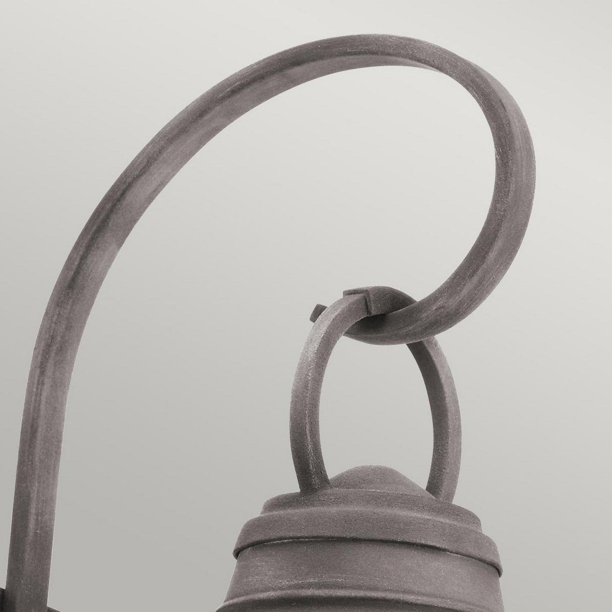 Close-up of the top portion of the Bridgepoint Outdoor 1 Light Medium Wall Lantern in weathered zinc. It showcases a curved metal arm with a loop design against a light background. This fixture has a rustic finish and includes seeded glass for added texture.