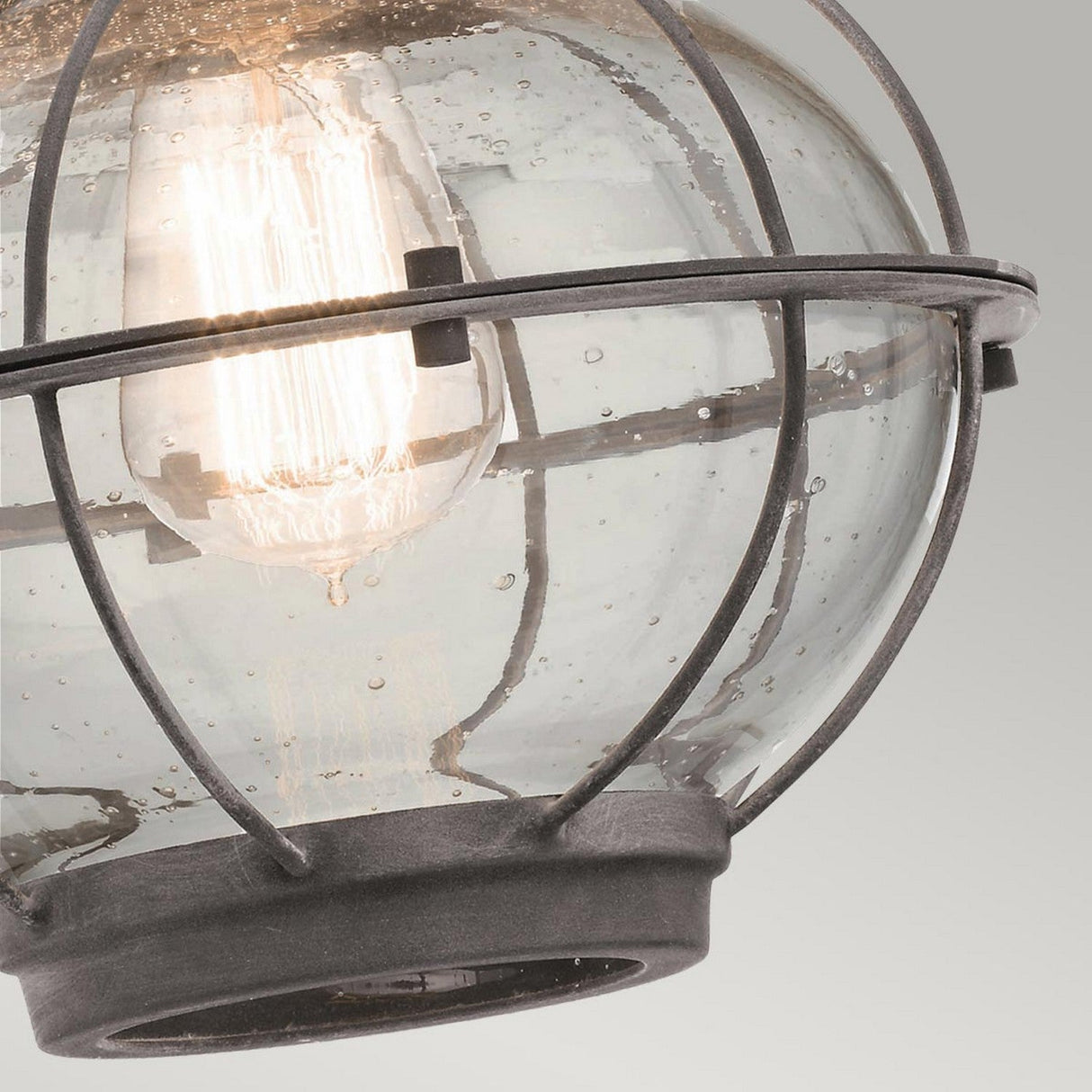 A detailed view of the Bridgepoint Outdoor 1 Light Medium Wall Lantern in Weathered Zinc showcases its vintage-style design, featuring a clear glass globe encased in a metal cage. The exposed bulb emits a warm glow against the neutral backdrop, enhancing its charming appearance.