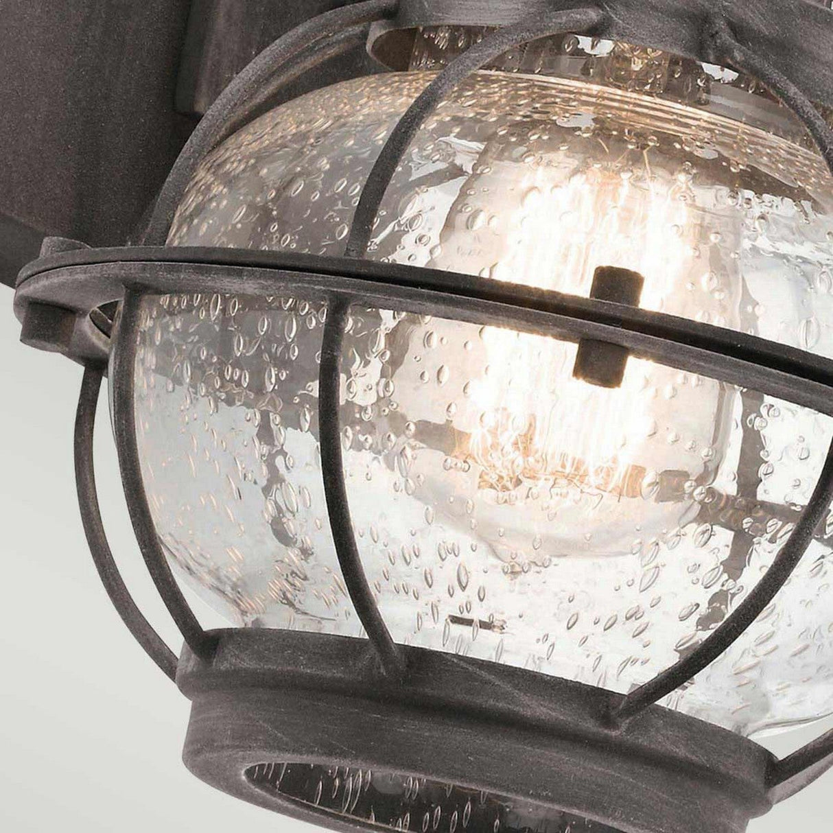 A close-up view of the Bridgepoint Outdoor 1 Light Small Wall Lantern reveals its rustic style, featuring a clear, rippled glass globe. The weathered zinc finish and metal guard give it a vintage appearance, while the bulb inside casts a warm glow.