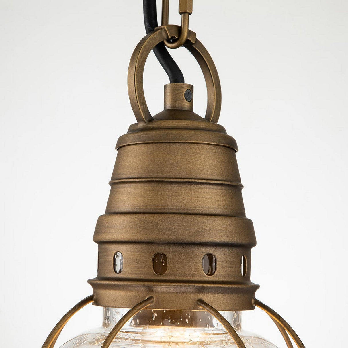 An up-close view of the Bridgepoint Outdoor 1 Light Small Chain Lantern showcases its vintage design with a natural brass finish. It features a cylindrical top adorned with circular cutouts and is complemented by a clear seeded glass shade, supported by metal brackets. The fixture is equipped with a black cord for easy hanging.