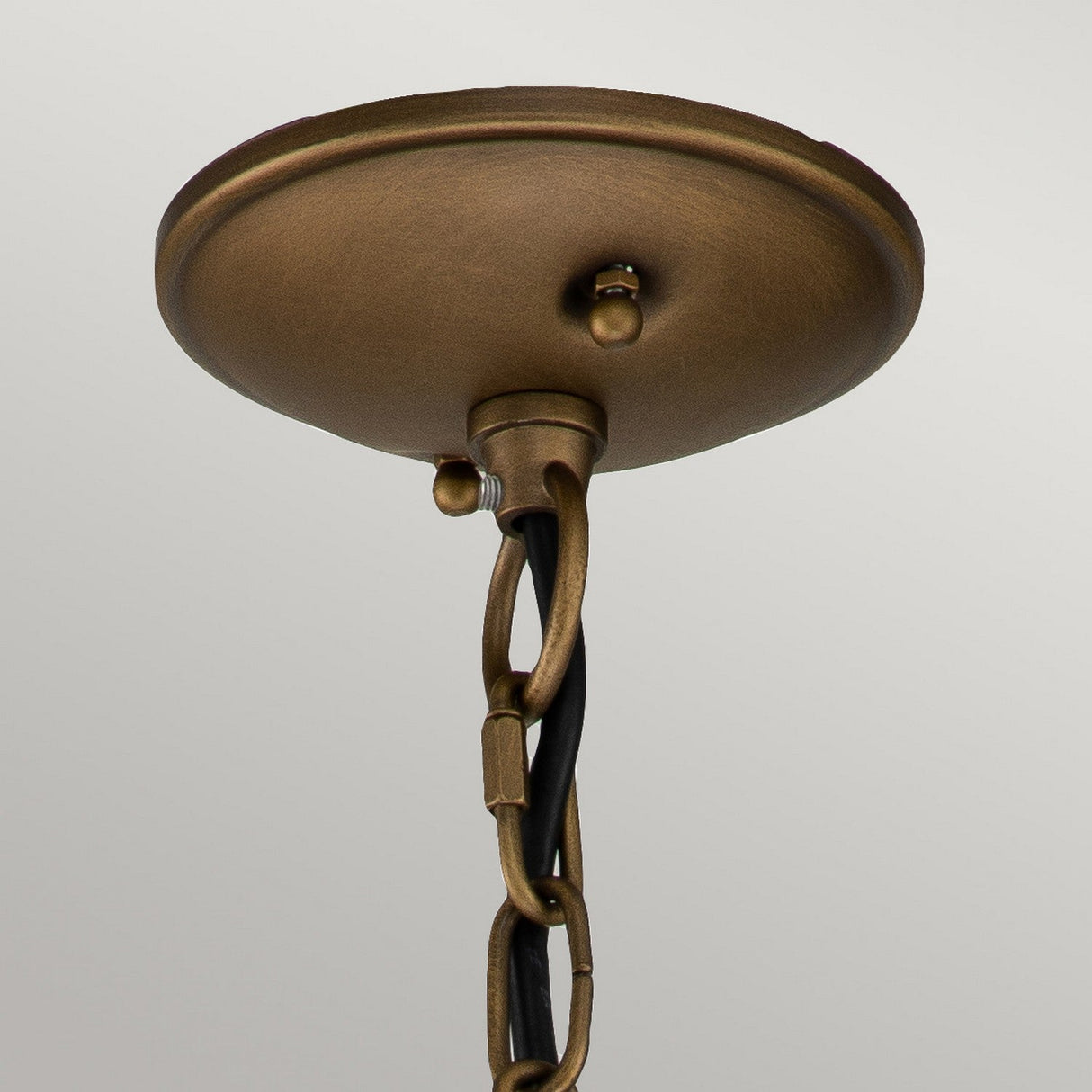 A detailed view of the Bridgepoint Outdoor 1 Light Small Chain Lantern - Natural Brass showcases its brass ceiling mount canopy with a black wire and chain. Set against a light gray background, this fixture's straightforward and industrial design is enhanced by its natural brass finish, echoing the charm of an outdoor lantern aesthetic.