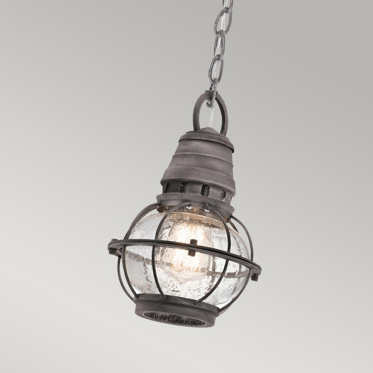 The Bridge Point Outdoor 1 Light Small Chain Light in Weathered Zinc exudes a vintage nautical charm with its weathered zinc metal frame and clear glass shade, suspended elegantly by a chain. The light inside is illuminated, highlighting the textured glass and casting a warm glow against the muted gray background.
