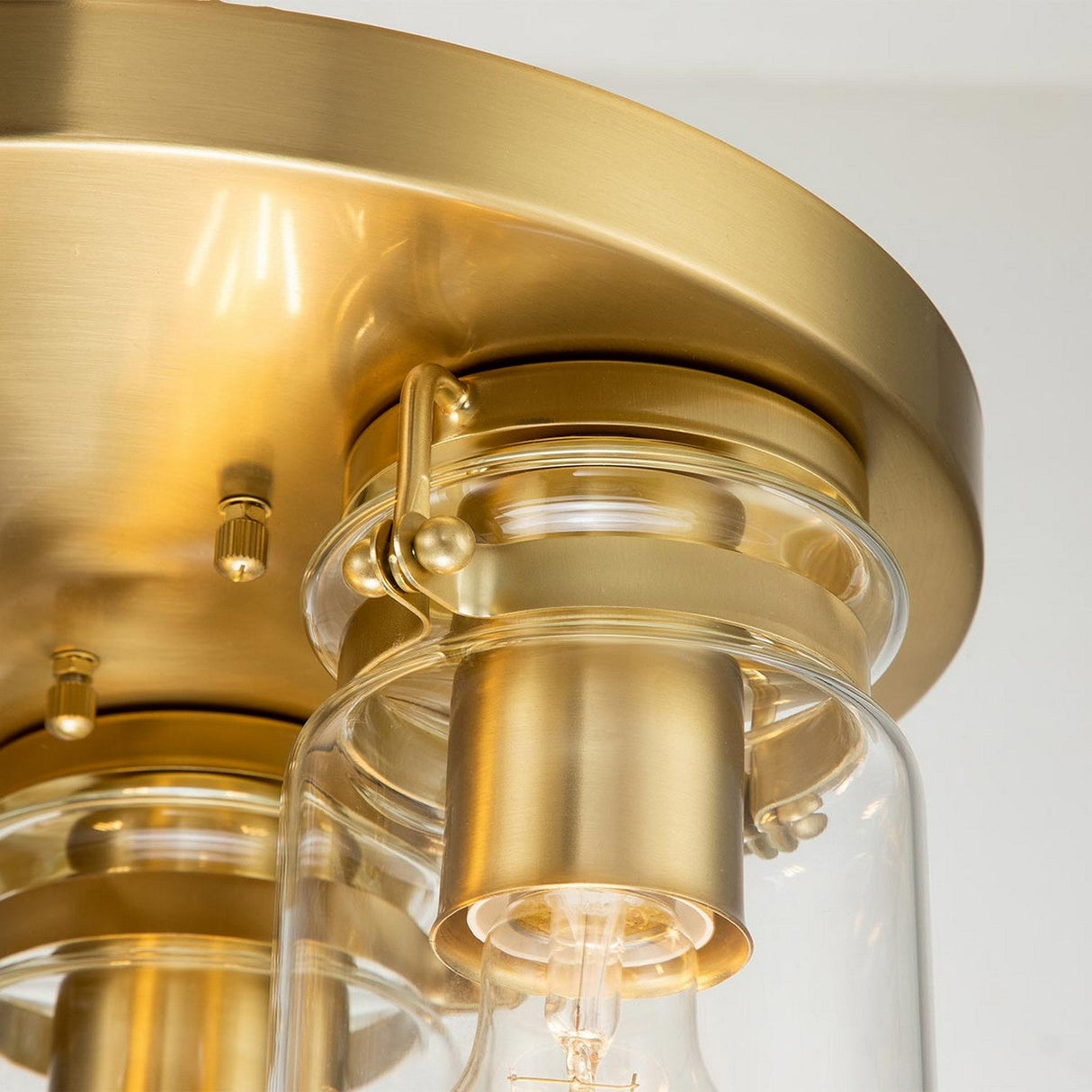 The Brinley 3 Light Flush - Brushed Brass ceiling fixture showcases a glass shade that allows the light bulb to be seen against a metallic backdrop, seamlessly blending vintage lighting aesthetics with a sleek, modern design.