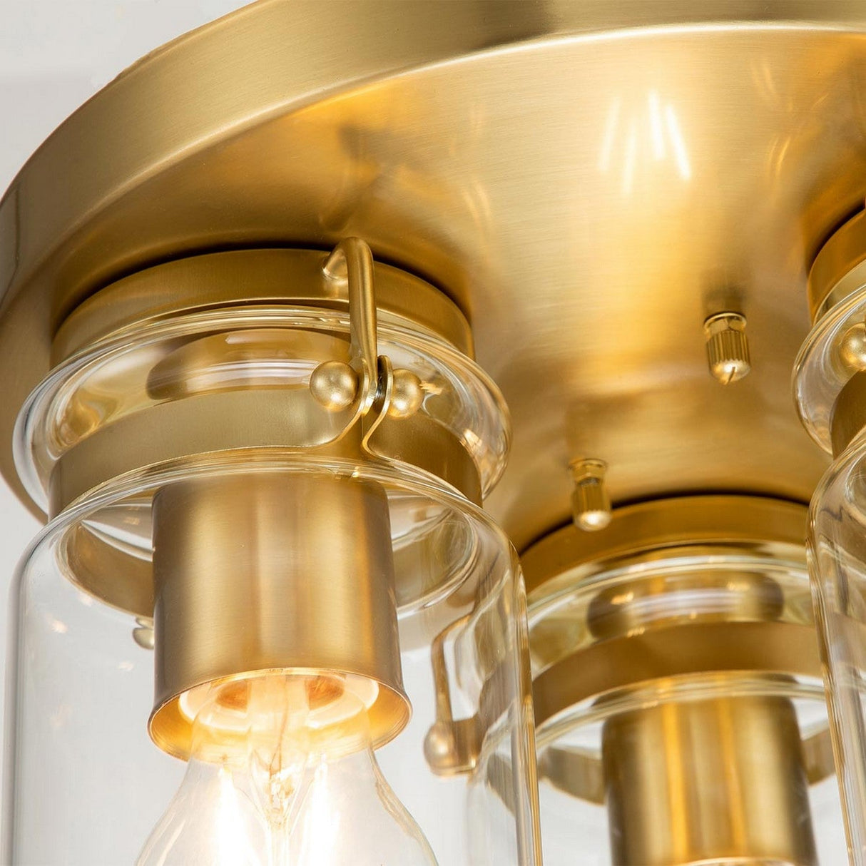 A close-up of the Brinley 3 Light Flush in Brushed Brass showcases its clear glass shades and vintage-inspired design. The exposed bulb and warm, reflective finish add an elegant touch to this modern ceiling fixture.