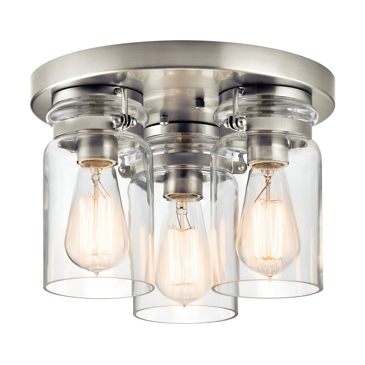 The Brinley 3 Light Flush in brushed nickel features a ceiling light fixture with three exposed Edison bulbs inside clear glass shades, mounted on a circular base. This vintage-inspired lighting creates a warm ambiance as the bulbs illuminate their surroundings.