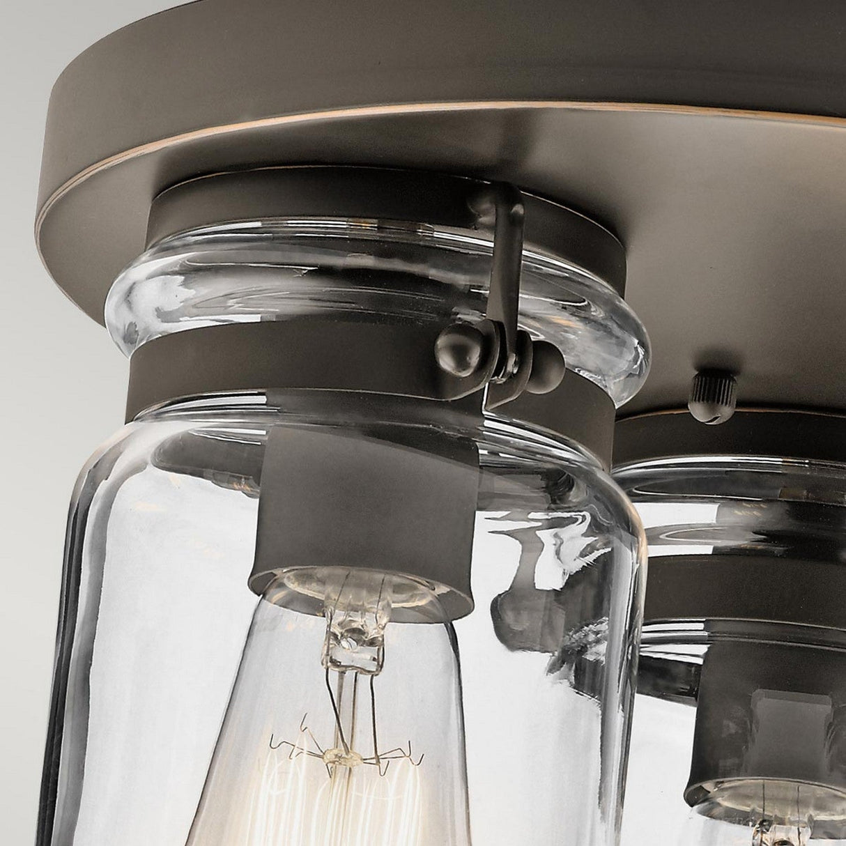 Close-up of the Brinley 3 Light Flush - Bronze ceiling fixture, highlighting its clear glass shades. Visible are two vintage-style Edison bulbs mounted on a round bronze base, exemplifying a combination of vintage-inspired and industrial design elements.