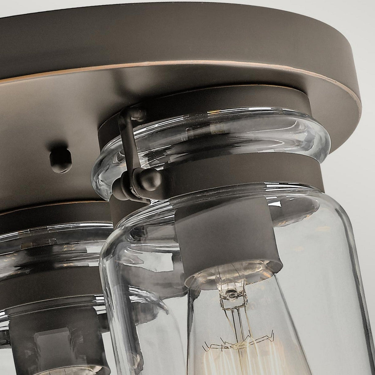 Close-up of the Brinley 3 Light Flush in bronze, showcasing its vintage-inspired design with clear glass dome covers that expose vintage-style filament bulbs. It is elegantly secured by a metallic band and screw, and mounted on a dark, circular ceiling plate.