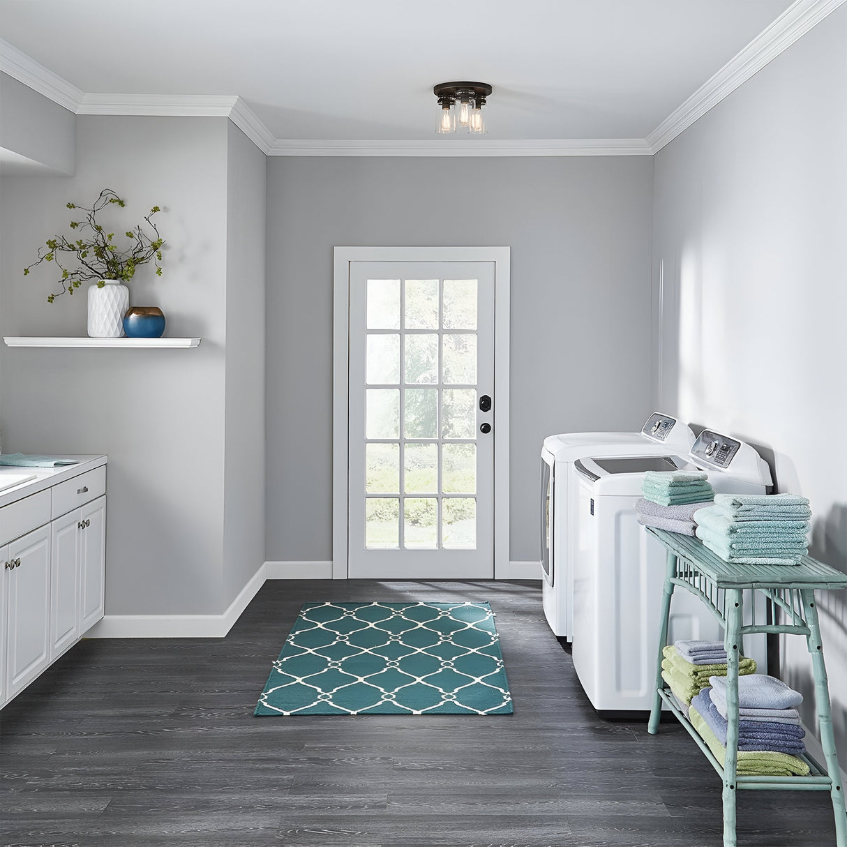 A modern laundry room with light gray walls and dark wood flooring showcases the Brinley 3 Light Flush in bronze, adorned with clear glass shades. The space includes a washing machine and dryer, a shelf displaying decorative vases, and an aqua and white patterned rug. Towels are neatly stacked on a green side table as natural light floods in through the glass door.