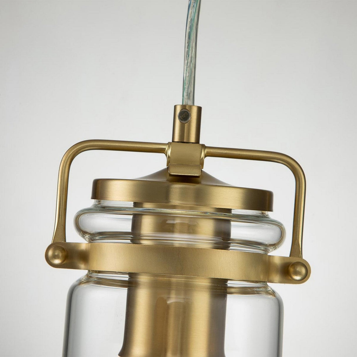 Close-up of the Brinley 1 Light Mini Pendant in a brushed brass finish, showcasing its vintage-style design with a transparent glass cover. The pendant features a cylindrical handle connected to the central brass structure, complemented by an exposed wire above.