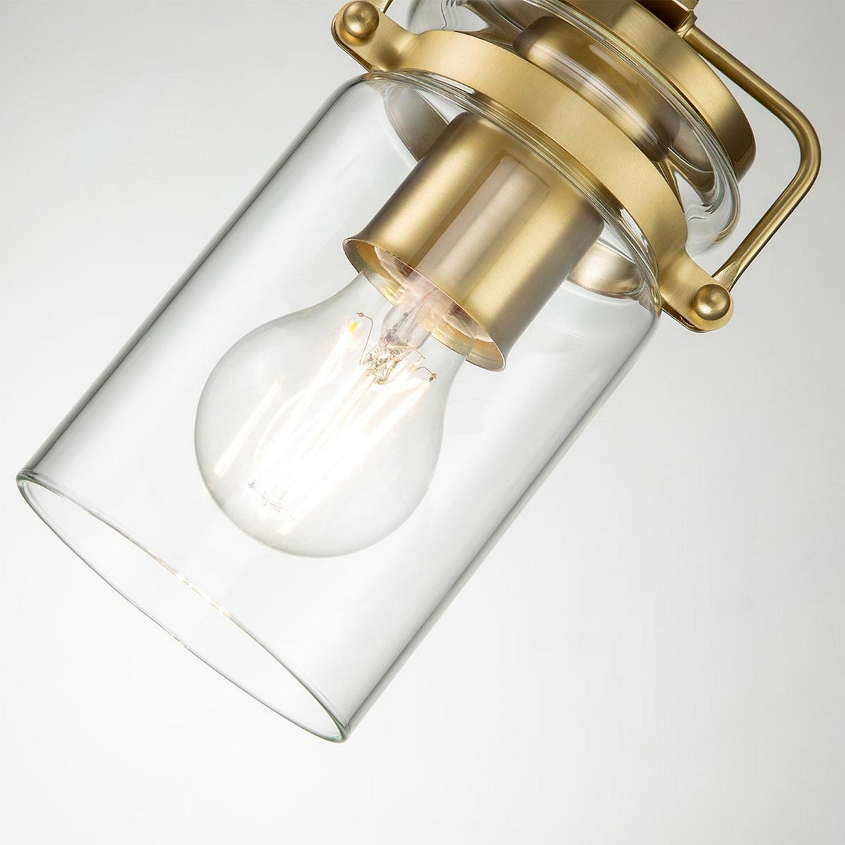 Close-up of the Brinley 1 Light Mini Pendant in brushed brass, featuring a clear glass shade. An exposed incandescent bulb inside emits a warm glow. The fixture is suspended against a neutral background.