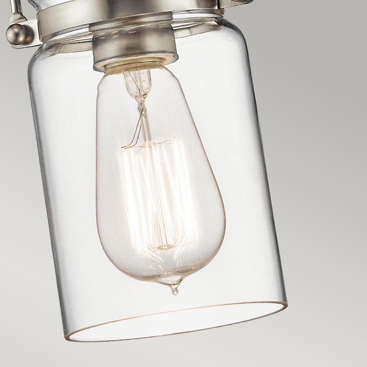 A detailed view of the Brinley 1 Light Mini Pendant - Brushed Nickel showcases a clear glass design with an exposed vintage-style Edison bulb. The bulb provides a warm glow, and the brushed nickel casing enhances its vintage-inspired charm against a neutral backdrop.
