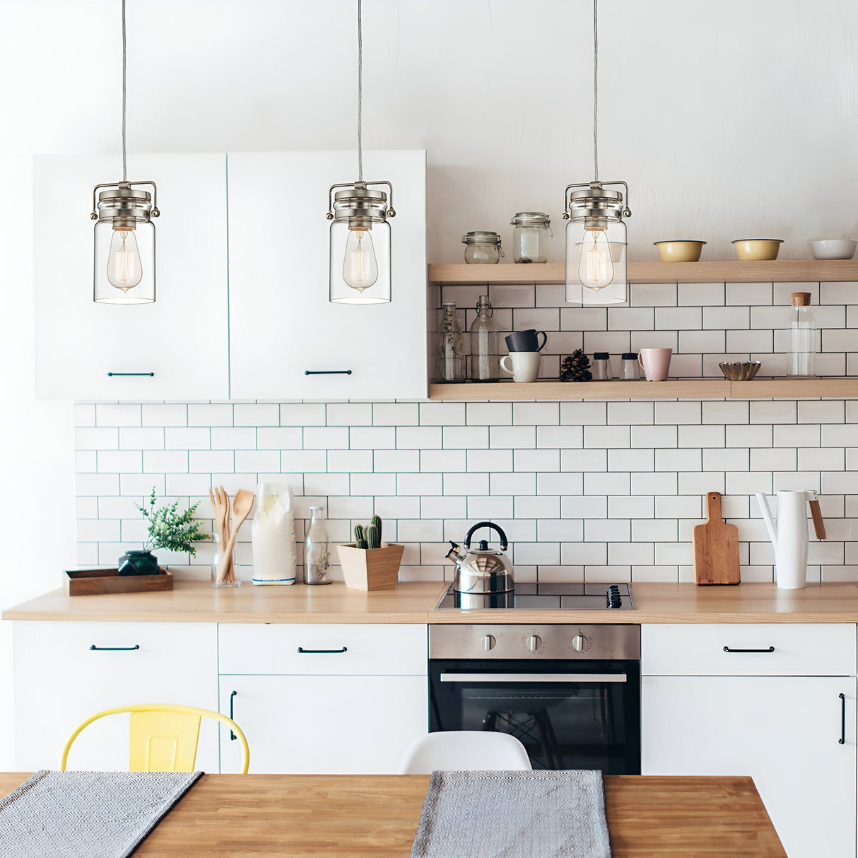 A modern kitchen showcases white cabinets and a wooden countertop paired with a tiled backsplash. Hanging above are three Brinley 1 Light Mini Pendants in brushed nickel, featuring clear glass shades. The counter is adorned with various kitchen items, plants, and a stovetop. Open shelves display bowls and jars, adding vintage-inspired charm to the space.