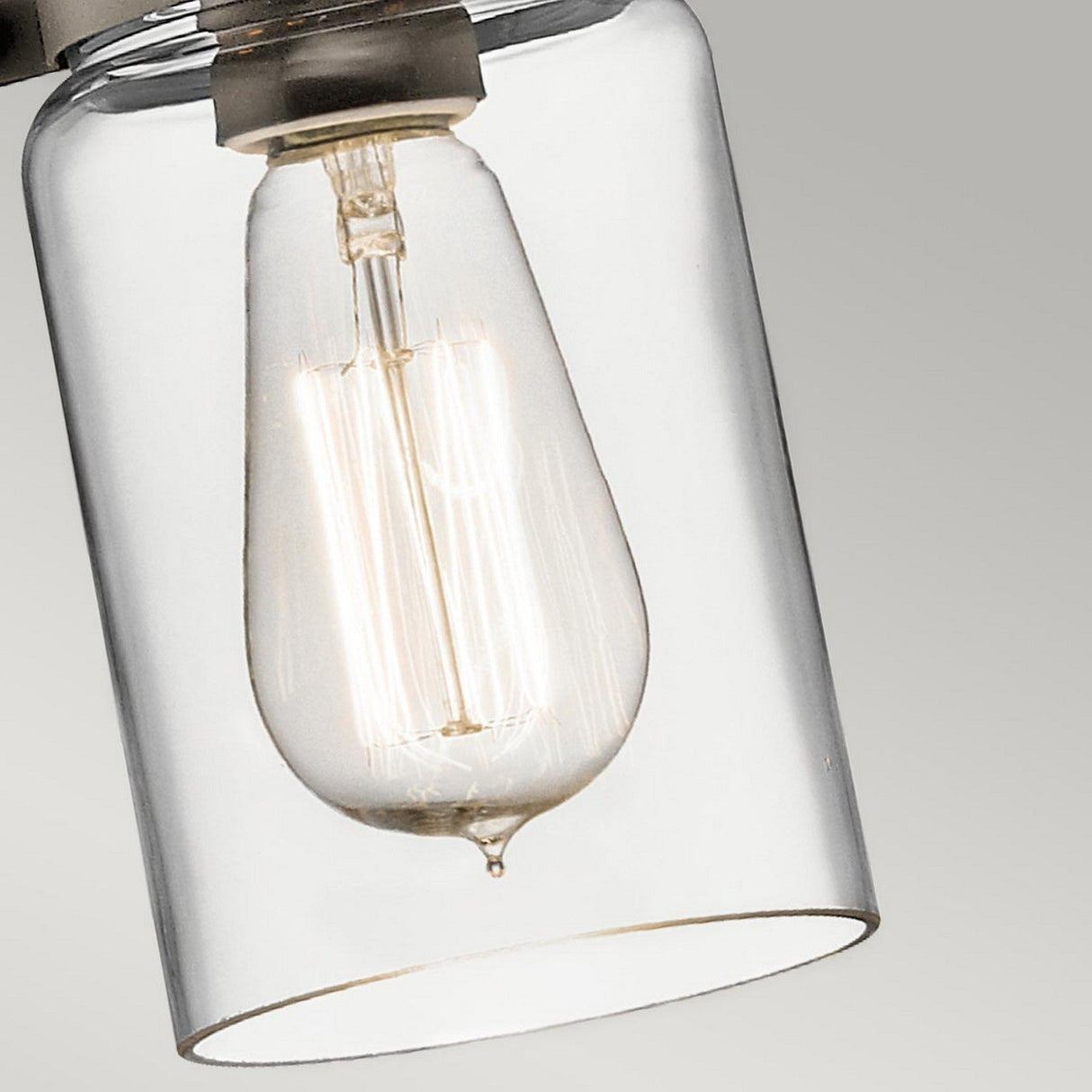 A close-up of the Brinley 1 Light Mini Pendant in bronze captures the Edison-style bulb glowing warmly inside its transparent glass cylindrical fixture. The backdrop of a plain gray complements rustic interiors, highlighting the fixture's understated elegance ideal for vintage lighting.