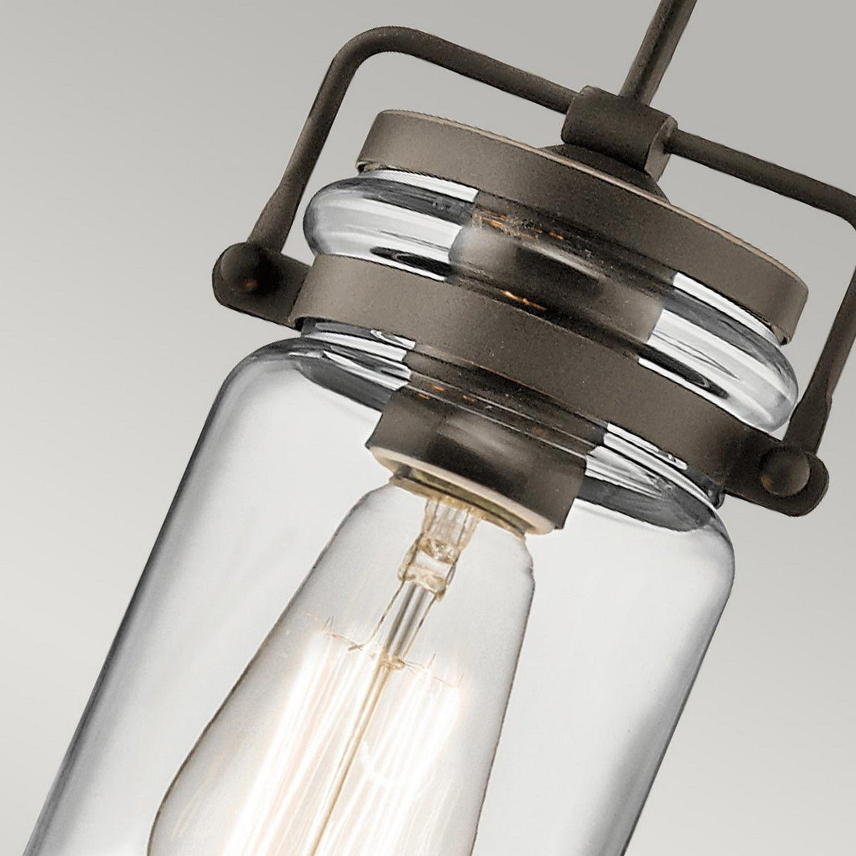 The Brinley 1 Light Mini Pendant - Bronze showcases a close-up of its vintage charm, featuring a clear glass encasing and a visible filament bulb. This fixture, ideal for rustic interiors, is supported by metal with an antique finish against a neutral background.