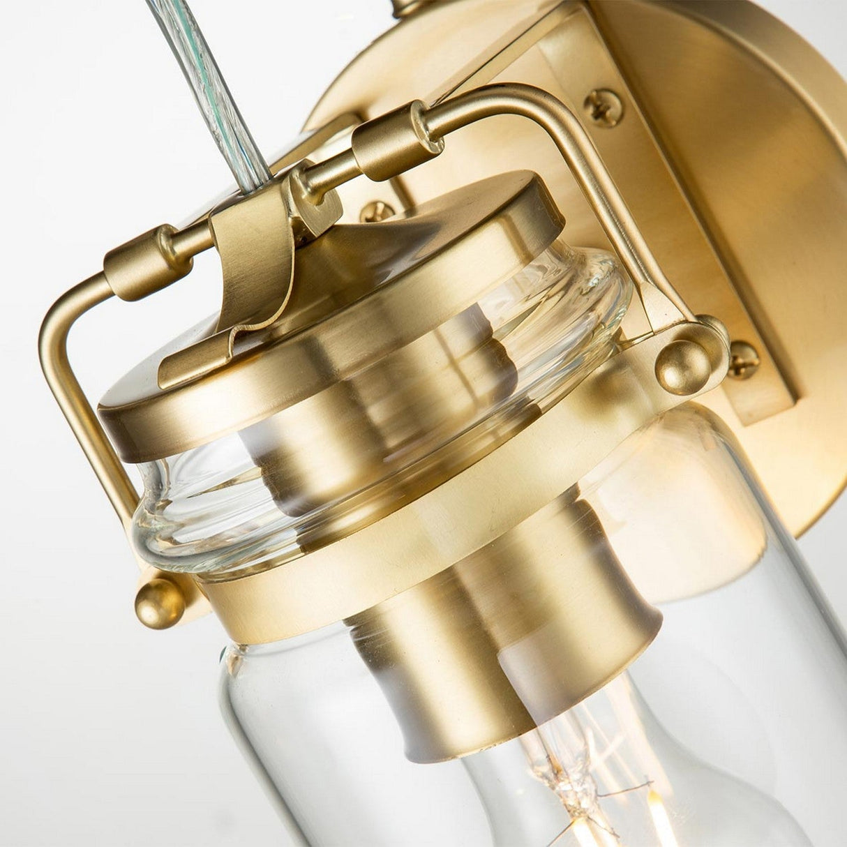 Close-up of the Brinley 1 Light Wall Light in brushed brass, showcasing a clear glass shade. Its cylindrical design with a visible light bulb emphasizes its modern industrial style complemented by vintage-inspired lighting elements.