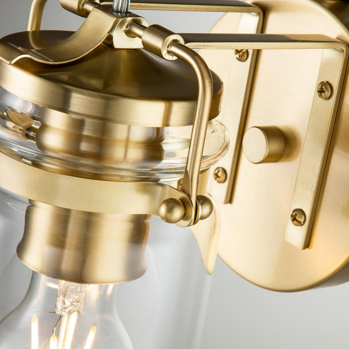 Close-up of the Brinley 1 Light Wall Light in brushed brass, featuring a clear glass cover. This design showcases elegant metallic accents and a visible lightbulb, highlighting its classic, vintage-inspired aesthetic.