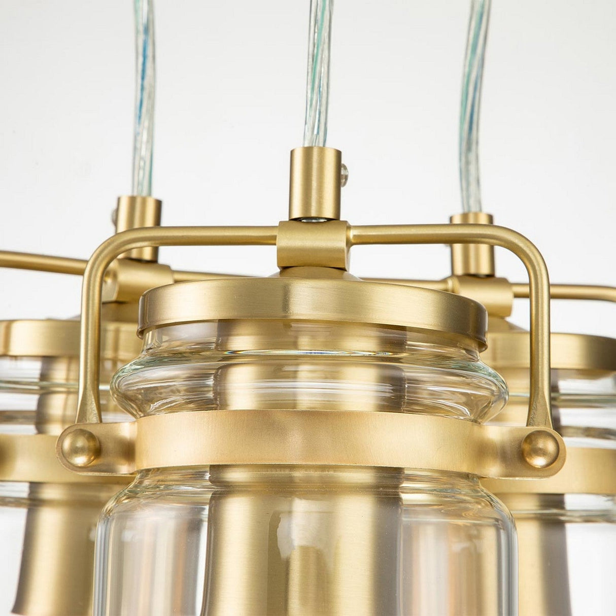 A close-up of the Brinley 3 Light Pendant - Brushed Brass highlights its cylindrical clear glass shades and brushed brass accents, complemented by sleek hanging cords, expertly blending industrial and contemporary design elements.