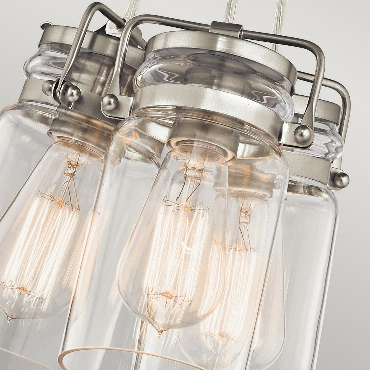 A close-up of the Brinley 3 Light Pendant - Brushed Nickel highlights its industrial-style design, featuring hanging lights with exposed filament bulbs encased in glass jars and complemented by brushed nickel fixtures. The bulbs cast a warm glow, accentuating the vintage aesthetic against a neutral background.
