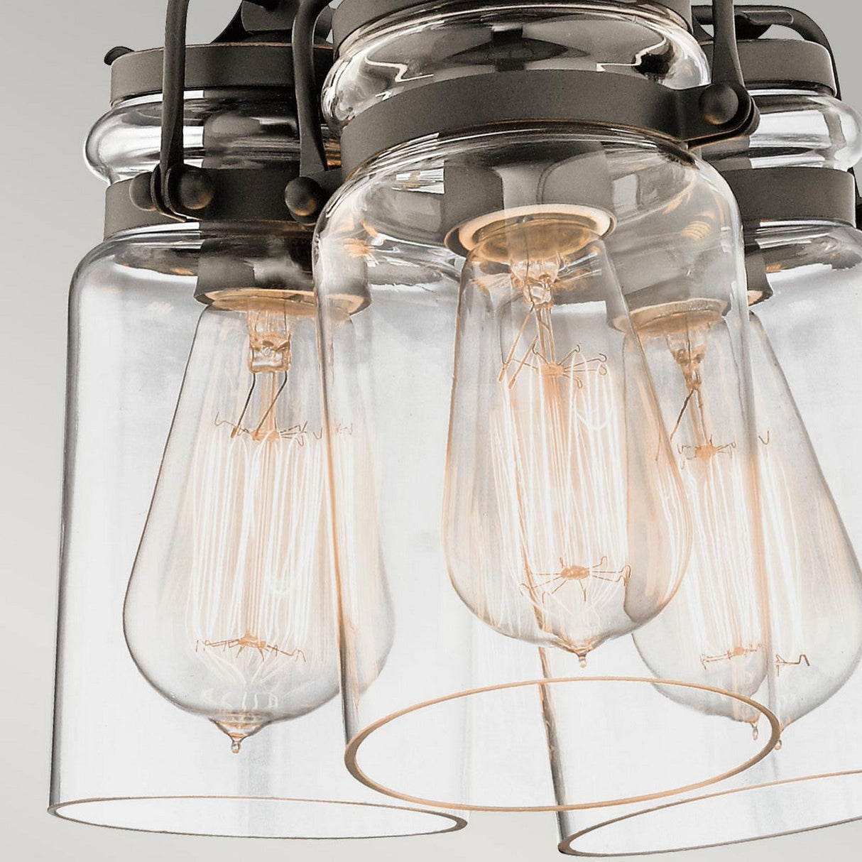 A close-up of the Brinley 3 Light Pendant in bronze showcases a modern industrial-style ceiling light with exposed vintage Edison bulbs encased in clear glass shades. The fixture exudes vintage charm, and its metal frame enhances the sleek, contemporary look, seamlessly blending old-world elegance with modern design.