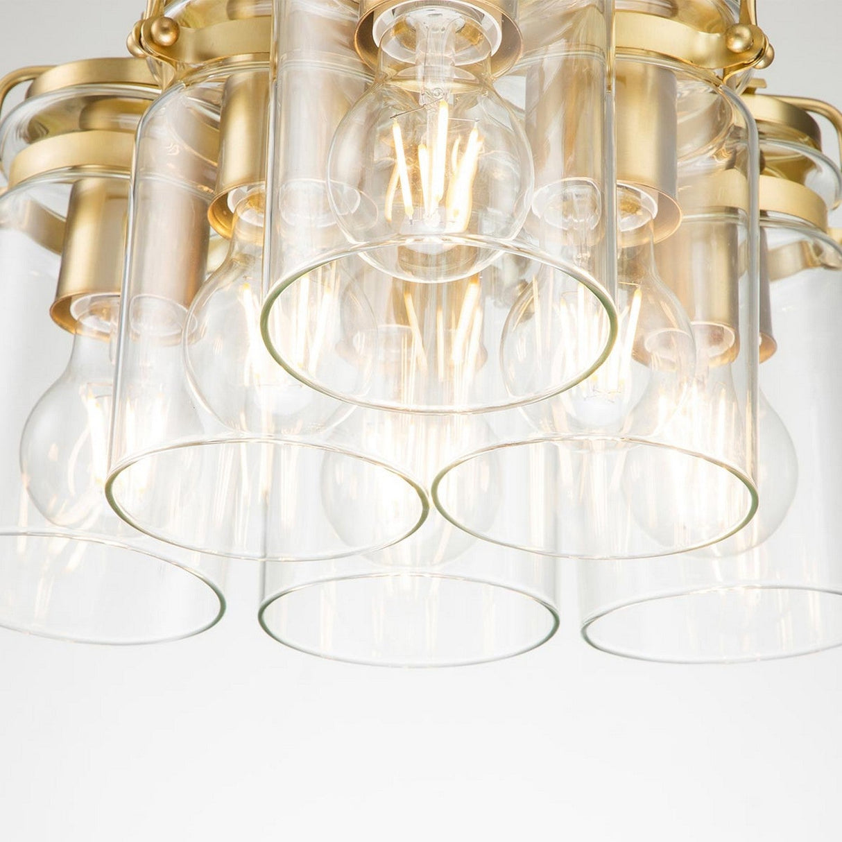 A close-up of the Brinley 6 Light Pendant showcases its exposed bulbs and cylindrical clear glass shades. The fixture's brushed brass pendant accents enhance its Mid-Century Modern vibe, featuring multiple light sources for a warm, inviting glow.