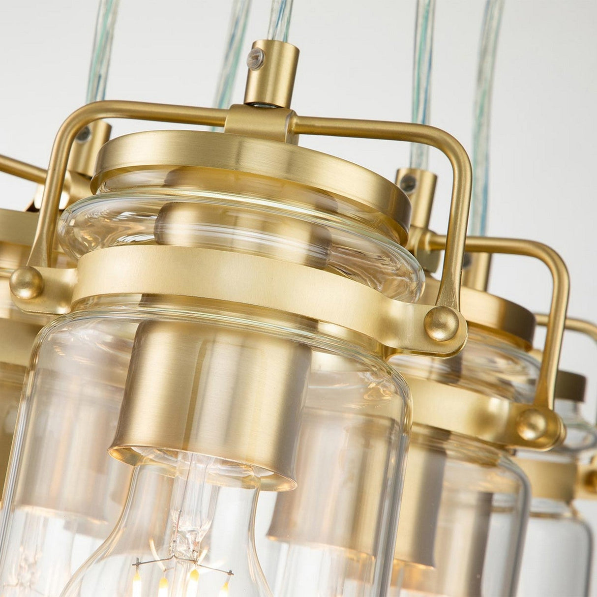 Close-up of three Brinley 6 Light Pendants in brushed brass, featuring clear glass surrounds and visible bulbs. This trio of lights is arranged to highlight the sleek, metallic design elements that echo a Mid-Century Modern style.