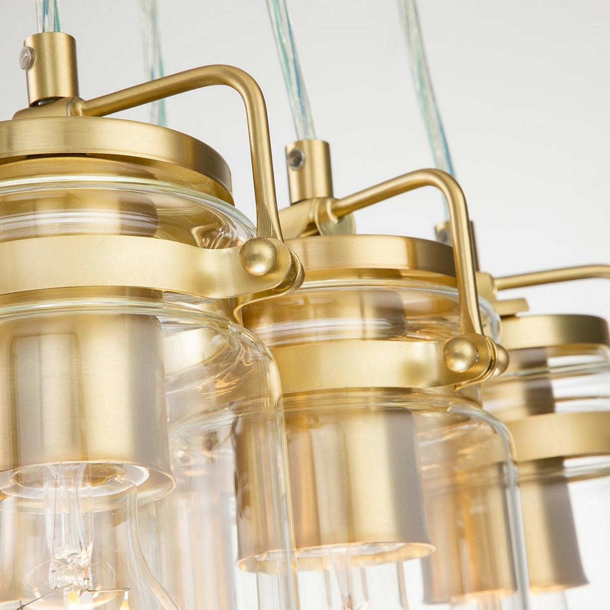 A close-up view of the Brinley 6 Light Pendant in brushed brass showcases three elegant pendant lights with clear glass shades, arranged in a row. This mid-century modern design features sleek fixtures suspended gracefully from cords, offering a timeless yet contemporary aesthetic.