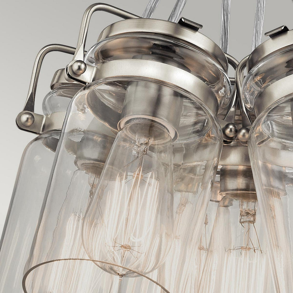 A close-up of the Brinley 6 Light Pendant in brushed nickel showcases several vintage-style light bulbs nestled within its design. Featuring exposed filaments and clear glass shades, this fixture exudes an industrial yet elegant aesthetic against a neutral background.