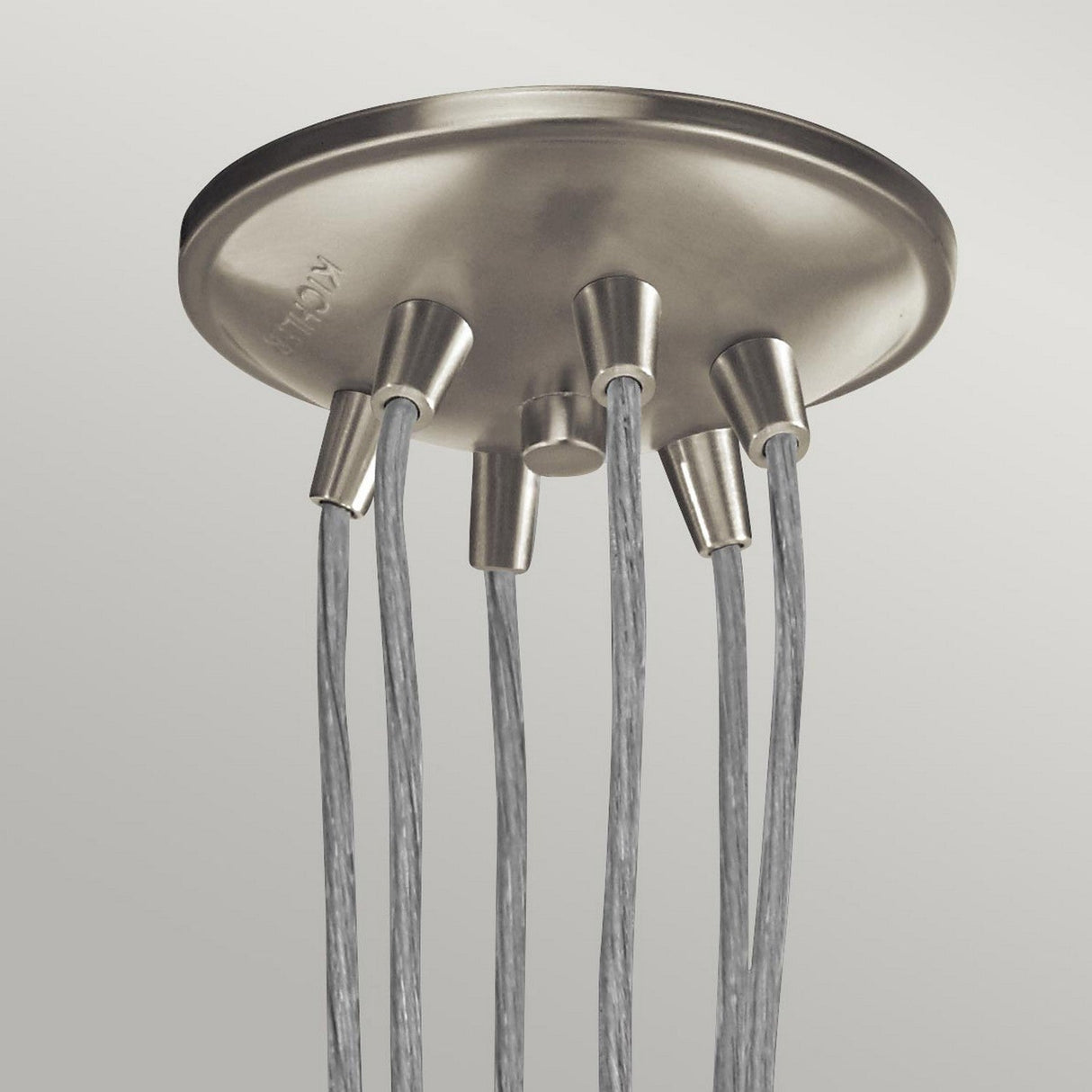 The Brinley 6 Light Pendant in brushed nickel showcases six suspended cables, each extending to a fixture adorned with clear glass shades, emanating from a central point. This design enhances a modern, minimalist aesthetic with vintage charm against a light gray backdrop.